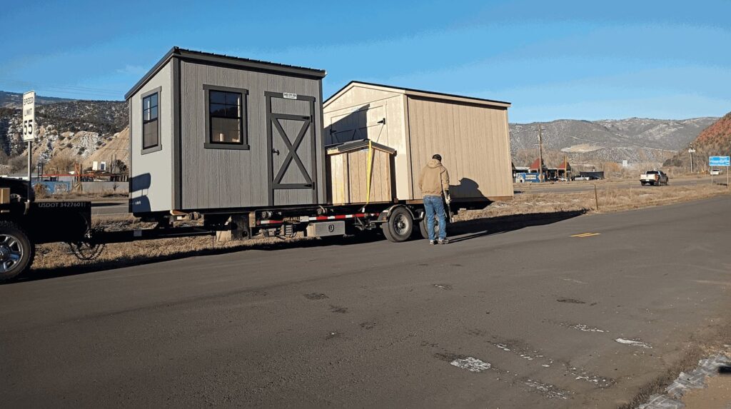 Two sheds and a smaller wooden structure strapped onto a flatbed trailer, parked beside a highway